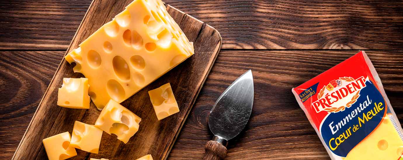Una cuña de queso emmental que tiene una parte cortada en daditos y desparramados sobre la madera en la que reposa y un pack de emmental Président